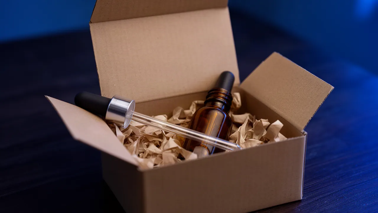 Een kartonnen verzenddoos met een bruin glazen flesje en een glazen pipet erin, op een donkere tafel